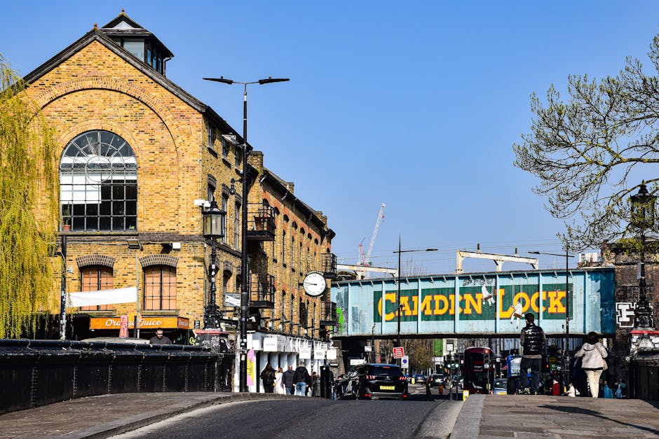 A street scene in Camden during daytime shows a brick building with large, semi-arched windows on the left side, adjacent to a busy pavement with pedestrians walking along. In the foreground, there are several cardboard boxes wrapped in plastic and fabric, as well as furniture pieces like a small table or cabinet, prepared for a home relocation. A wheeled trolley is visible near the boxes, used for transporting household items, while a van is parked close by, suggesting ongoing furniture transport or loading. To the right, a large blue shipping container with the words 'Camden Lock' painted in yellow is elevated on a bridge structure spanning the street, indicating the area's association with market and delivery activity. The background features a clear blue sky, with some trees and construction cranes visible further along the street, emphasizing the urban environment typical of Camden's lively market and moving logistics scene. Camden Movers are likely involved in the packing and moving process depicted here, supporting efficient transportation of household goods during market day moves.