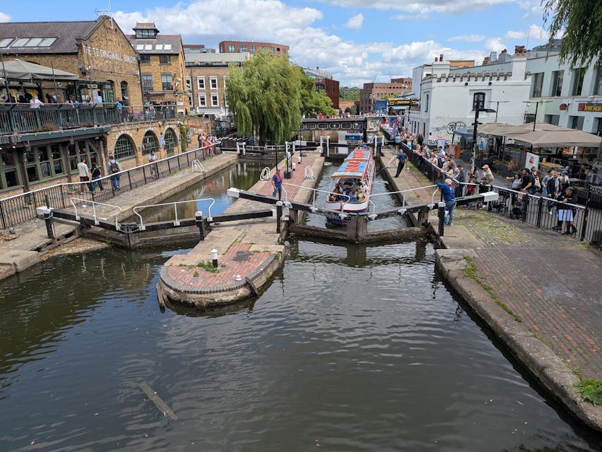 A canal scene in Camden with a narrowboat passing through a lock, surrounded by people walking along the pathways and standing by the railings. On the right side, there are outdoor seating areas and shops, with some individuals appearing to assist in the loading process by carrying boxes and furniture wrapped in plastic and blankets. The lock gate is open, and a person is seen operating it with a control lever. The area features brick pathways, metal railings, and a lush green tree providing shade. The background shows a mix of modern and historic buildings under a partly cloudy sky. Camden Movers' van or trucks are not visible, but the scene reflects typical elements involved in home relocation and furniture transport in a busy urban setting, suitable for a house removals service specializing in packing and moving logistics during market day moves in Camden.