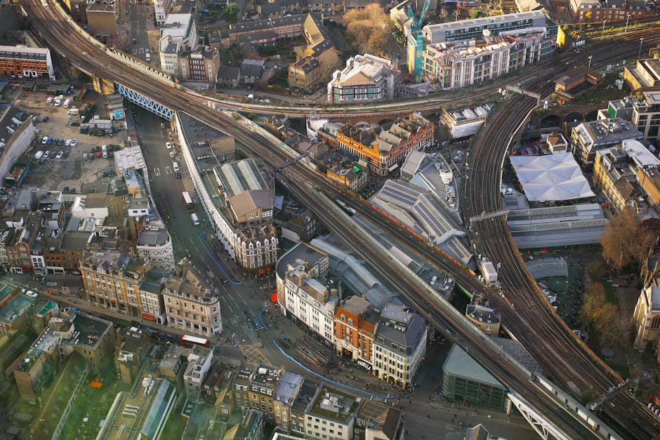 An aerial view of a busy urban area with a complex network of roads, bridges, and railway tracks crossing each other. Several small commercial buildings and residential structures occupy the streets below. A white van is visible parked on a pavement near a small street, while nearby, a trolley and moving blankets suggest a furniture transport process associated with house removals. The scene includes materials such as cardboard boxes and plastic wrap ready for home relocation inside the property or for transport. Camden Movers, a professional removals company, is likely involved in the loading and packing activities, taking advantage of a narrow street entrance to facilitate efficient furniture transport and moving logistics. The lighting indicates late afternoon with warm sunlight casting shadows across the scene, capturing the detailed movements of a house removal in an urban environment, suitable for a page about removal advice for narrow streets like Chalk Farm Road.