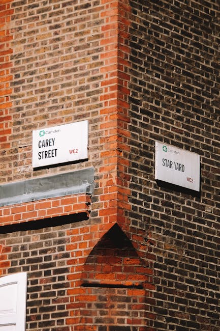 Close-up of a corner where two brick walls meet, each displaying street signs for Carey Street and Star Yard, both with the Camden logo and mention of WC2 postcode. The Carey Street sign is mounted above a horizontal vent or grille with a metallic cover, while the Star Yard sign is positioned lower, aligned with the brickwork. The brickwork features varying shades of red and brown, with some mortar lines visible, and the lighting highlights the textures of the bricks and signs. This setting illustrates typical urban street signage in Camden, relevant for home relocation and moving services, such as those provided by Camden Movers, especially when planning furniture transport along narrow streets.