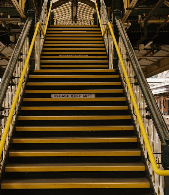 A set of outdoor pedestrian stairs with black steps marked by a central yellow line, metal handrails on both sides, leading up to a covered footbridge structure with a sloped roof. Surrounding the sta